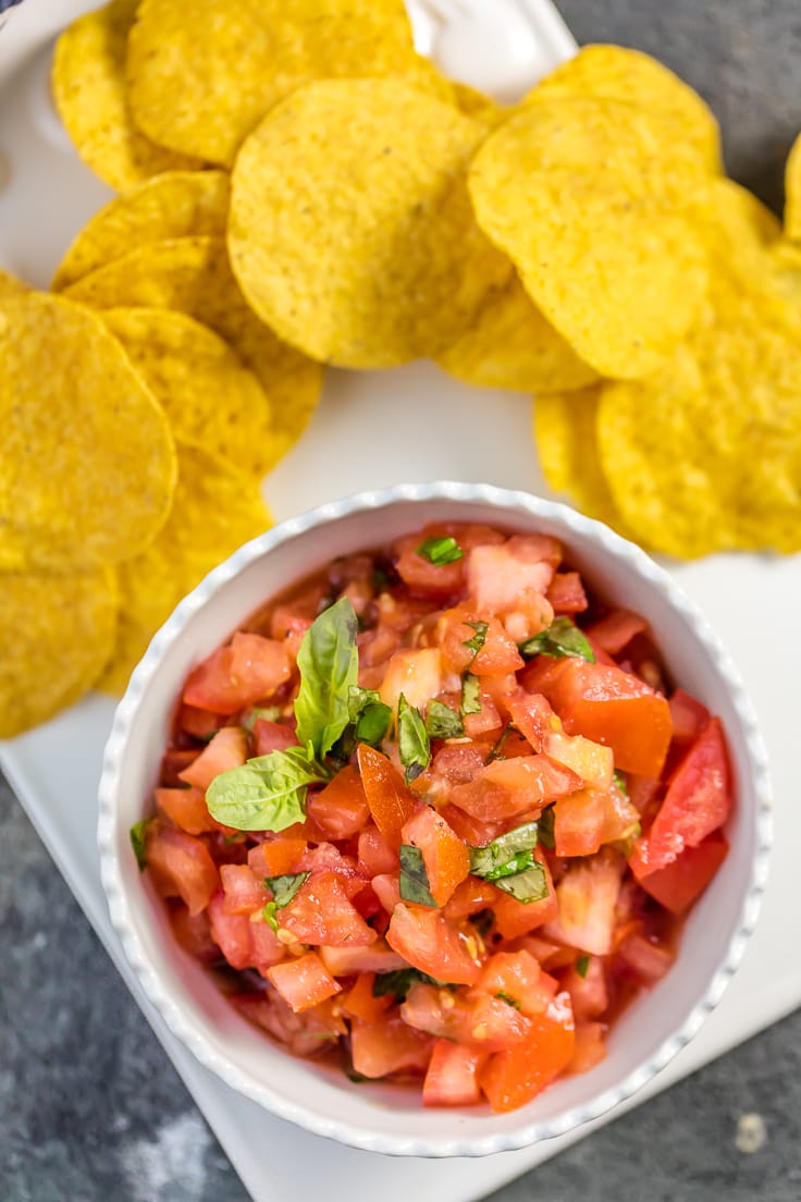 White bowl of Italian salsa with tortilla chips on white plate.