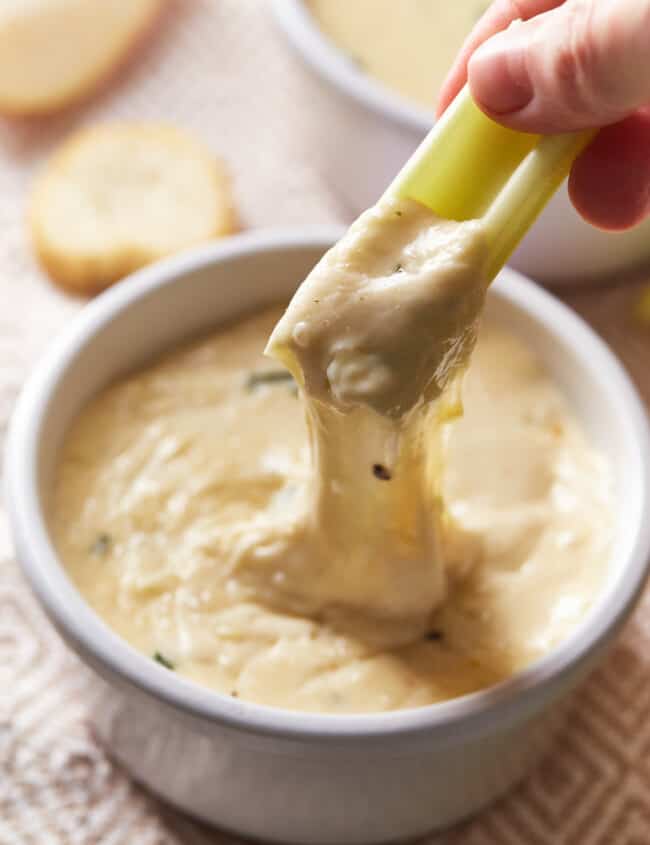 a hand pulling a piece of celery out of white wine fondue in a white bowl, creating a cheese pull.