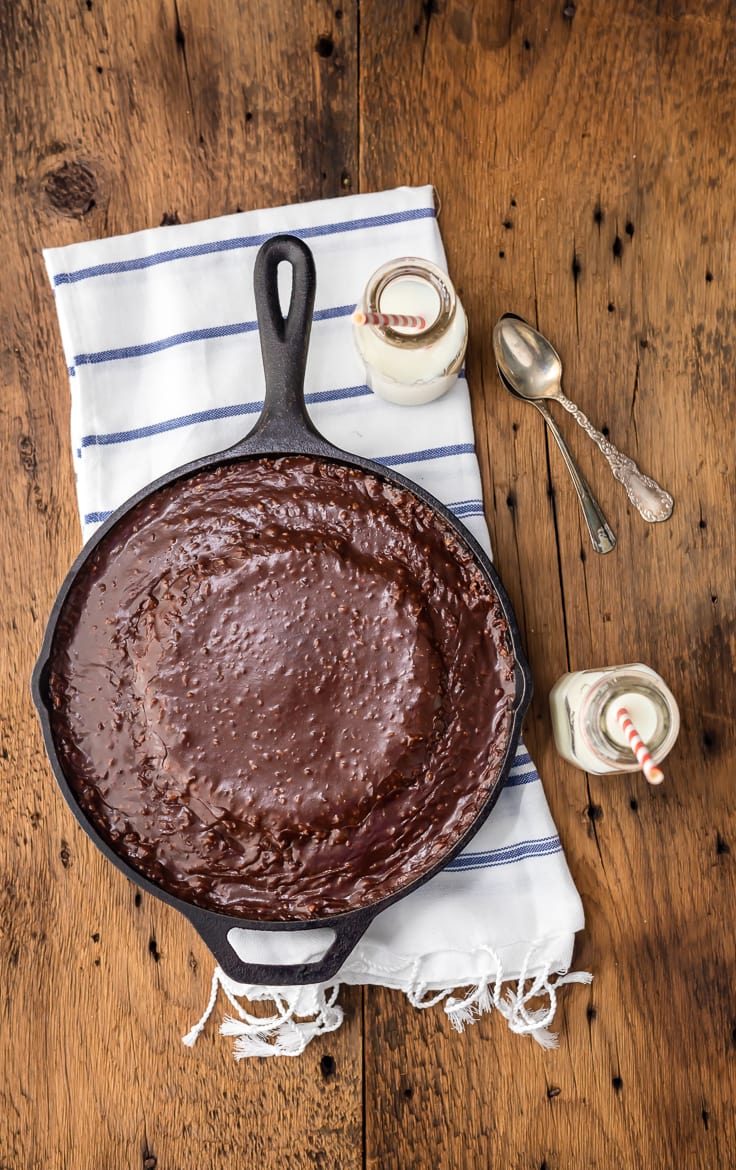 a skillet filled with chocolate cake sitting on a striped dish towel, with two small jars of milk and two spoons