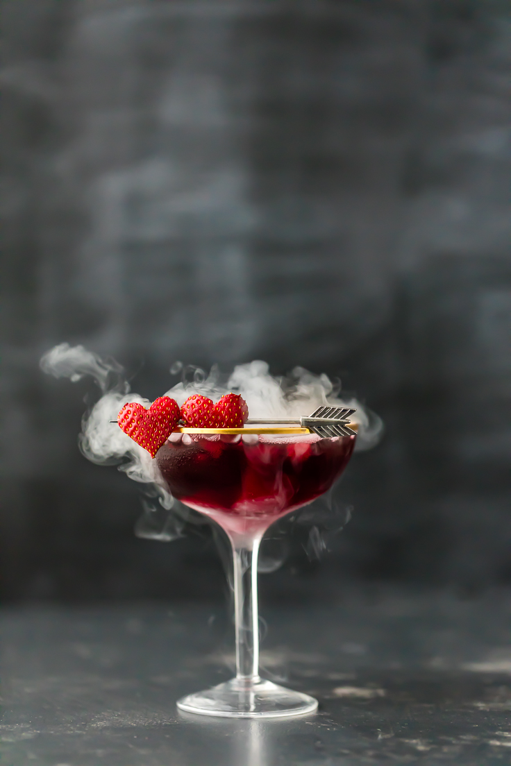 a purple martini in a glass with heart shaped strawberries and dry ice