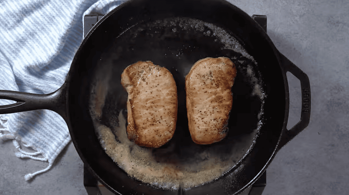 Two pork chops are being cooked in a skillet with apple butter.