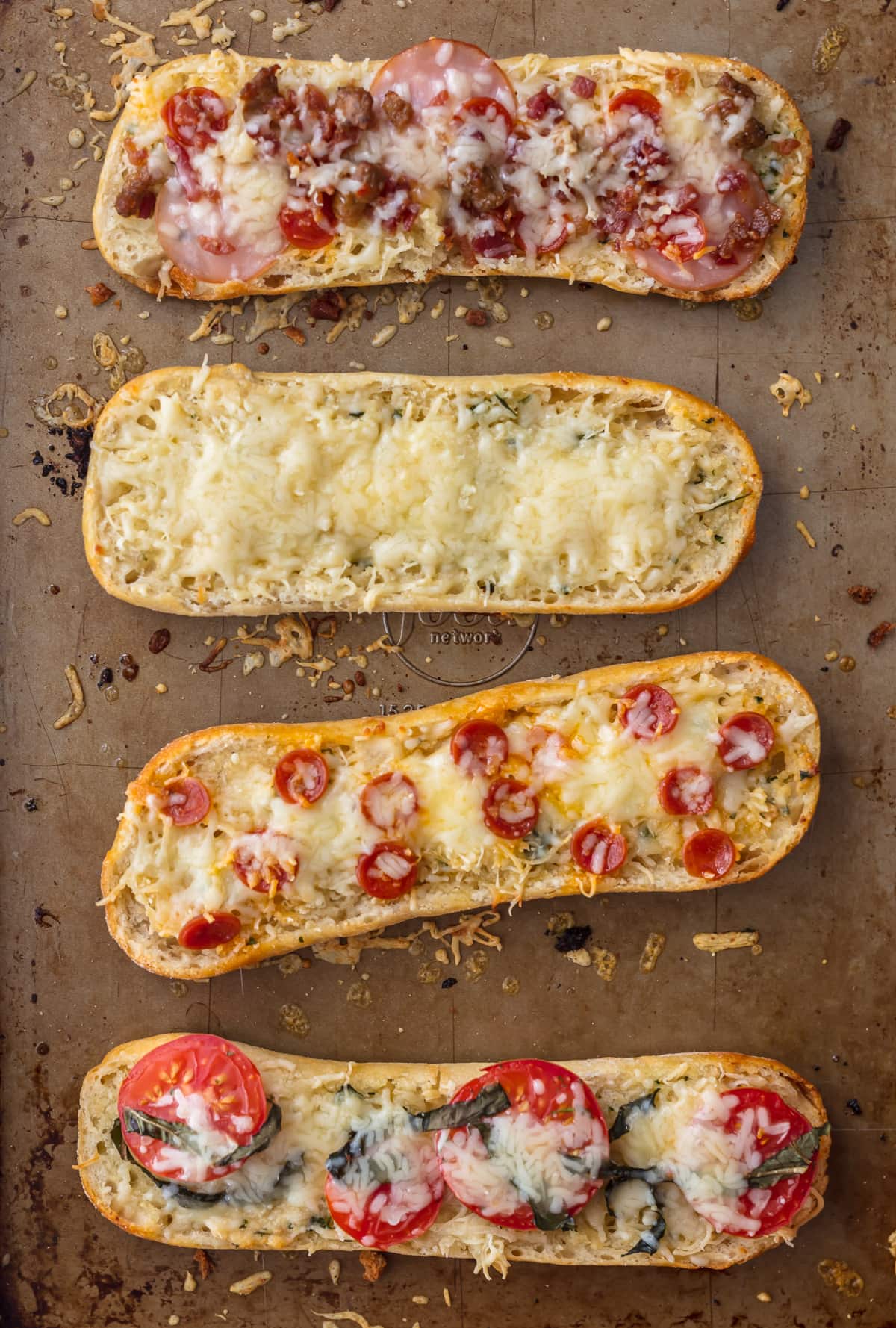 Four french bread pizza on a baking sheet: one topped with meat, one with cheese, one with pepperonis, one with tomatoes and basil