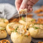 a hand scooping dip out of a bread bowl with a pretzel