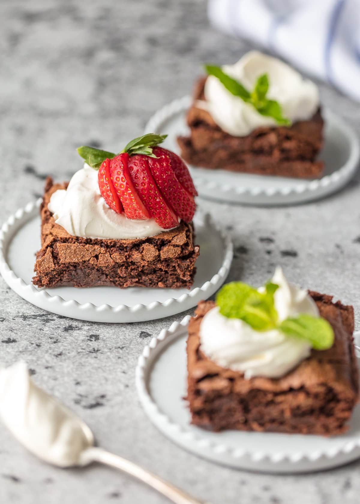 sliced homemade brownie from scratch on a white plate topped with whipped cream, berries, and mint.