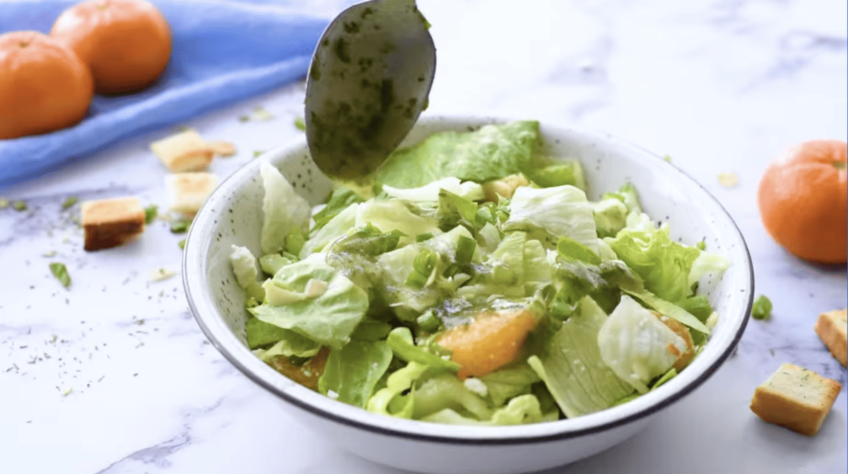 a spoon drizzling cider vinaigrette over mandarin orange salad in a white bowl.