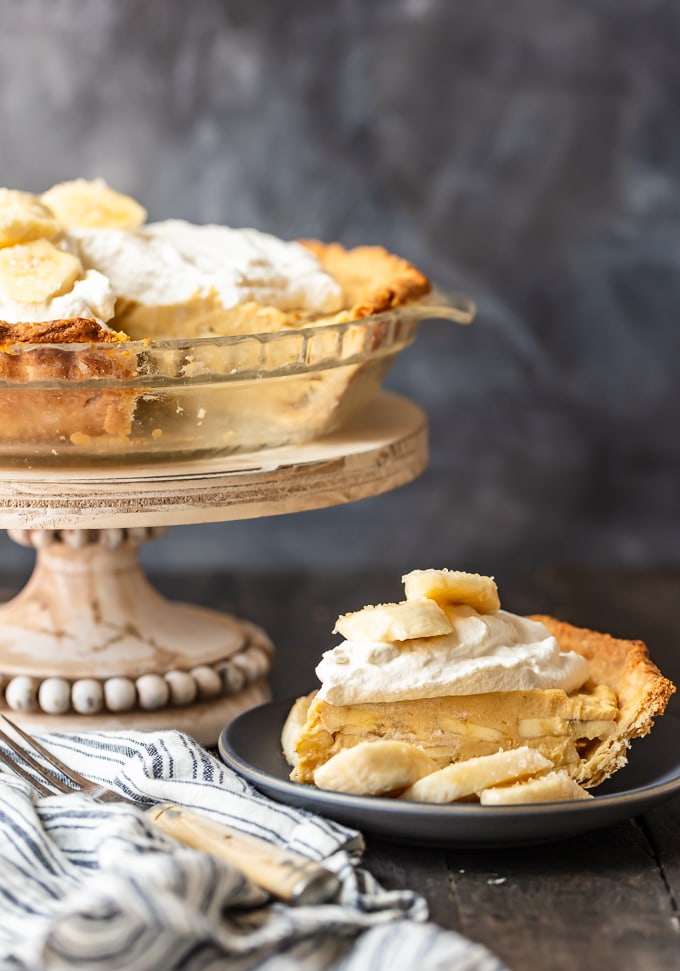 A full banana cream pie on a pedestal, next to a plate of a slice of pie