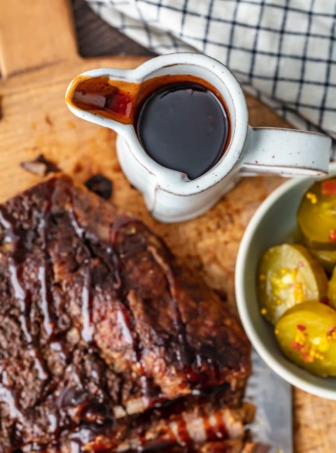 a container of brisket sauce next to a bowl of pickles and a piece of brisket