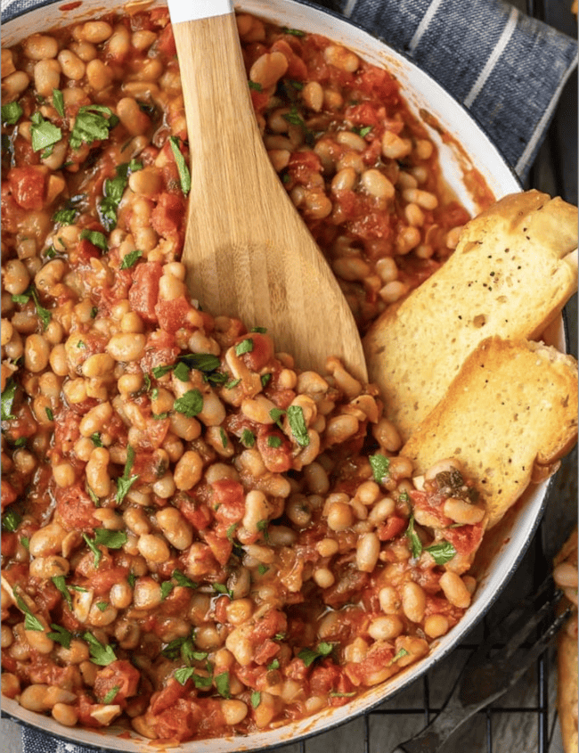 A pan full of white beans in tomato sauce, being stirred with a wooden spoon.