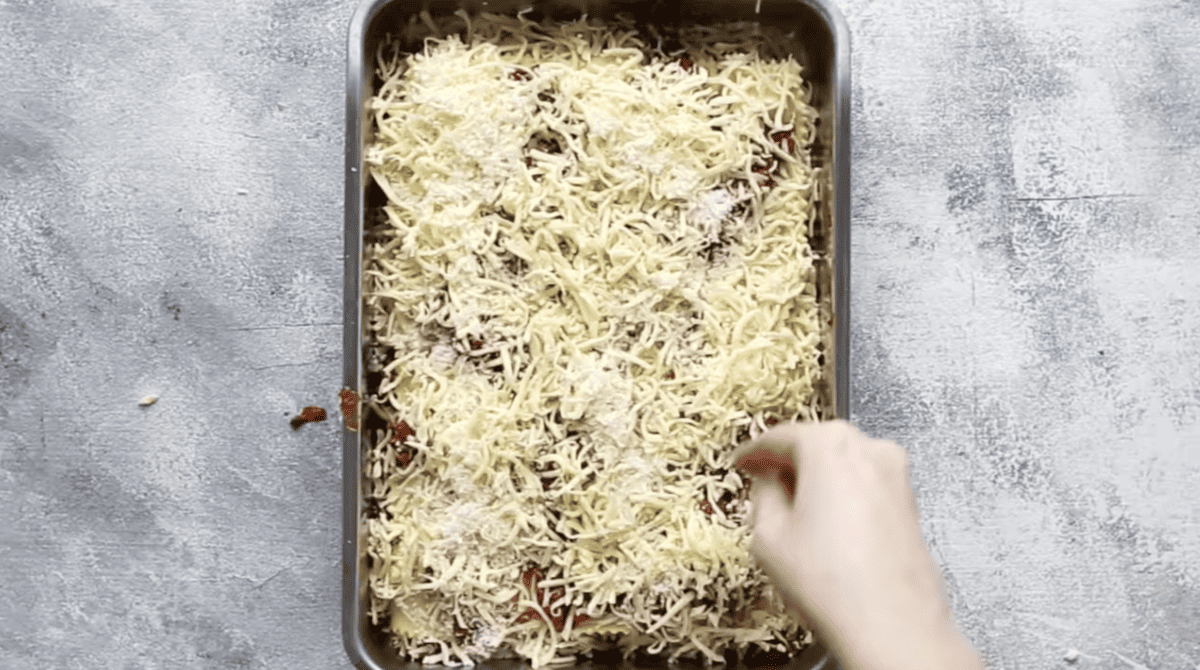 A person putting cheese into a lasagna baking pan.