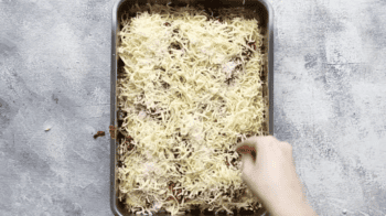 A person putting cheese into a lasagna baking pan.