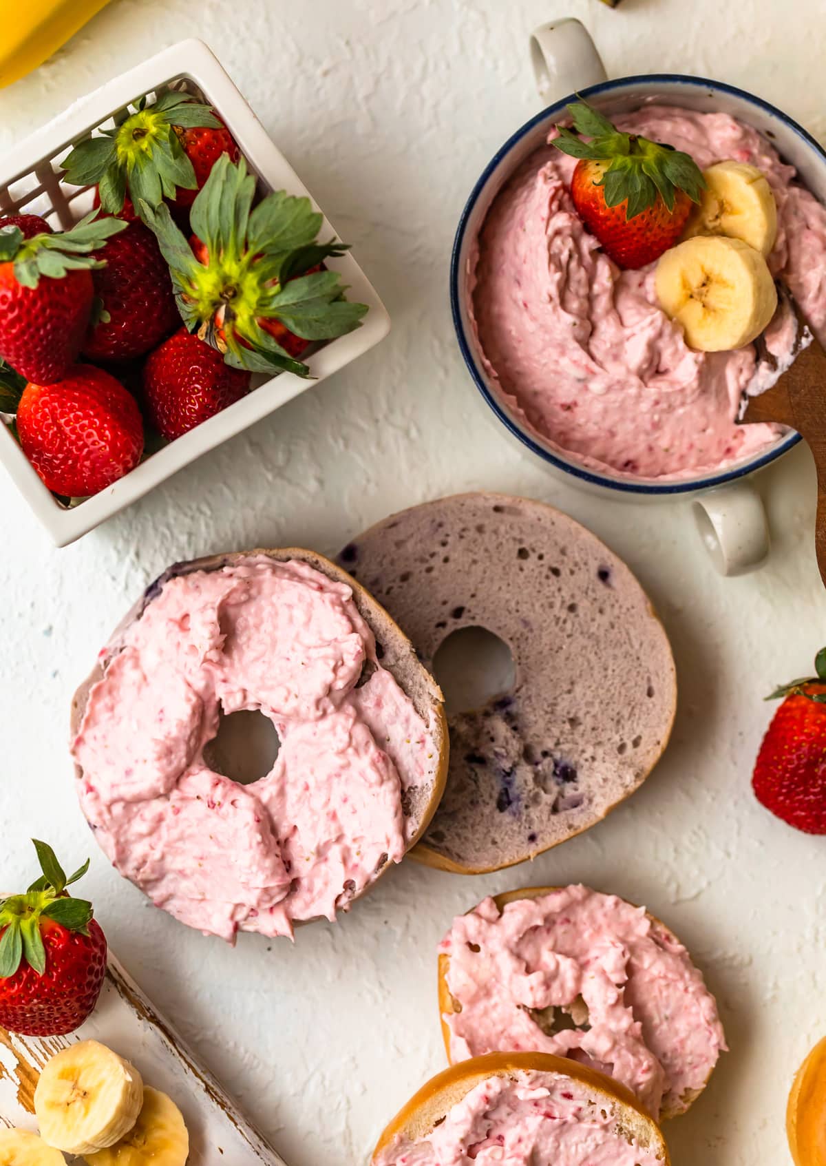 a bagel with cream cheese next to a basket of strawberries and a bowl of strawberry cream cheese