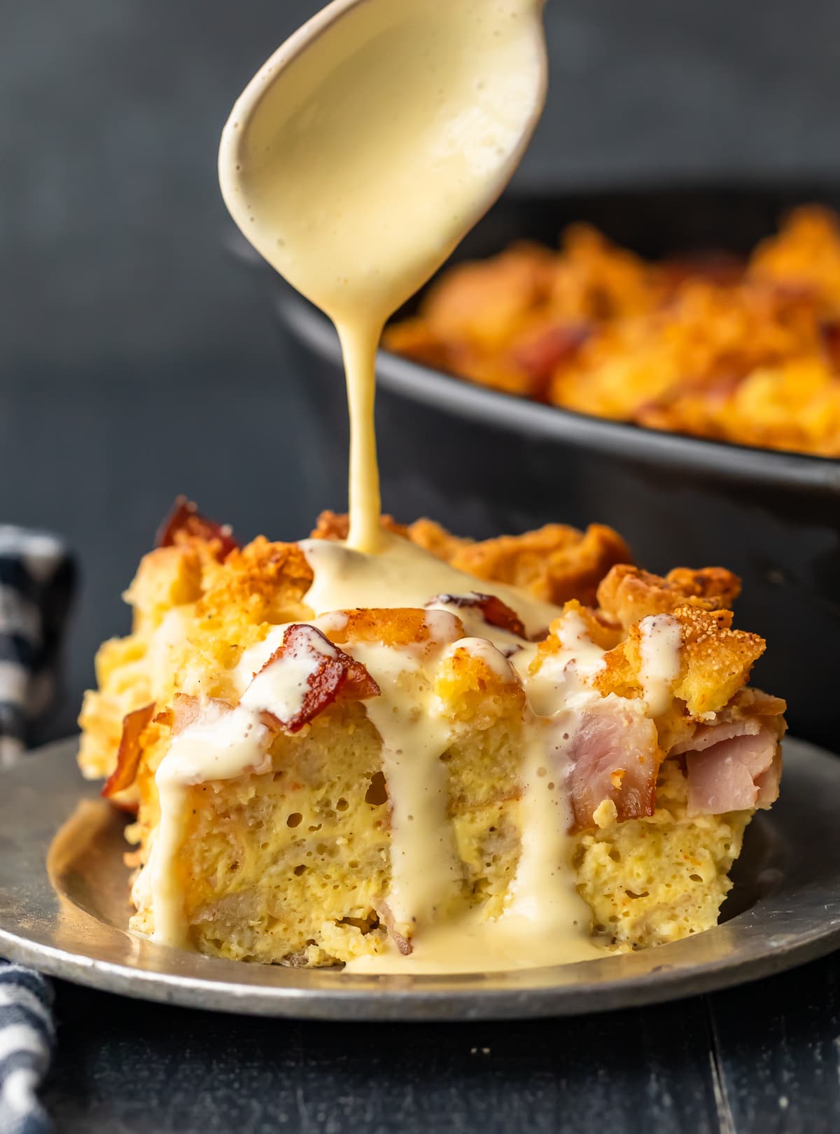 spoon pouring hollandaise sauce over a slice of eggs benedict casserole.