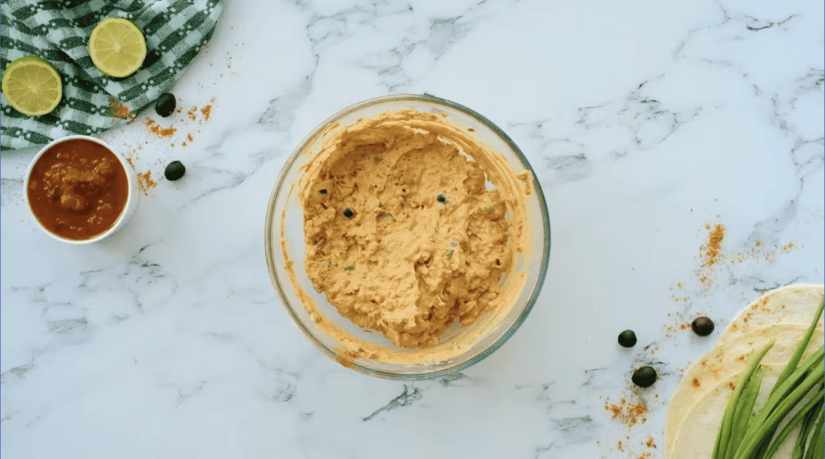 A bowl of guacamole and tortilla chips on a marble table next to Mexican pinwheels.