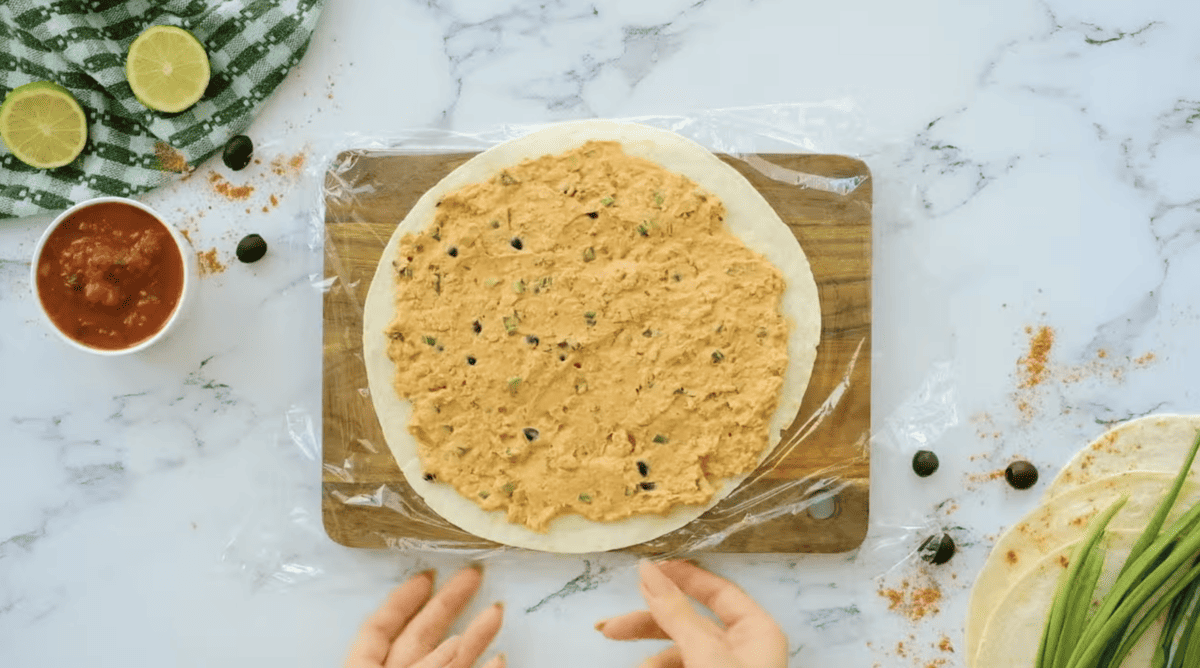 A person is preparing a tortilla for sausage pinwheels on a cutting board.