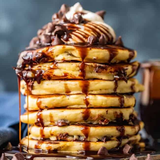 lots of chocolate chip pancakes stacked on each other drizzled with chocolate syrup and whipped cream