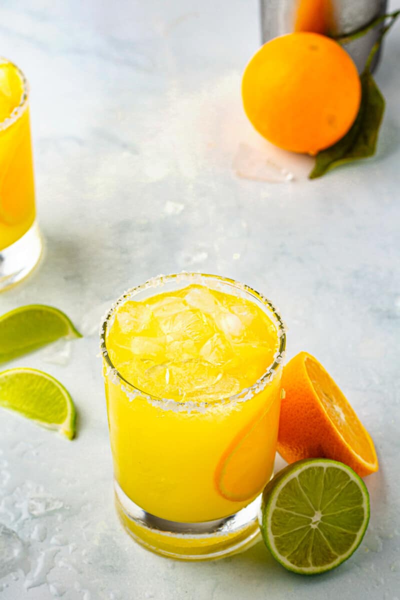 orange margarita in glass next to limes and oranges