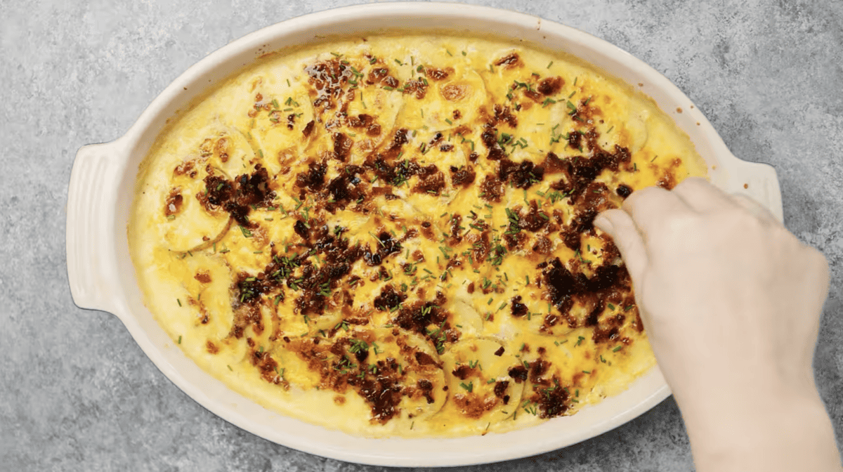 A person reaches into a dish of loaded scalloped potatoes.