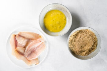 tilapia filets on a white plate, egg mixture in a white bowl, and breading mixture in a white bowl