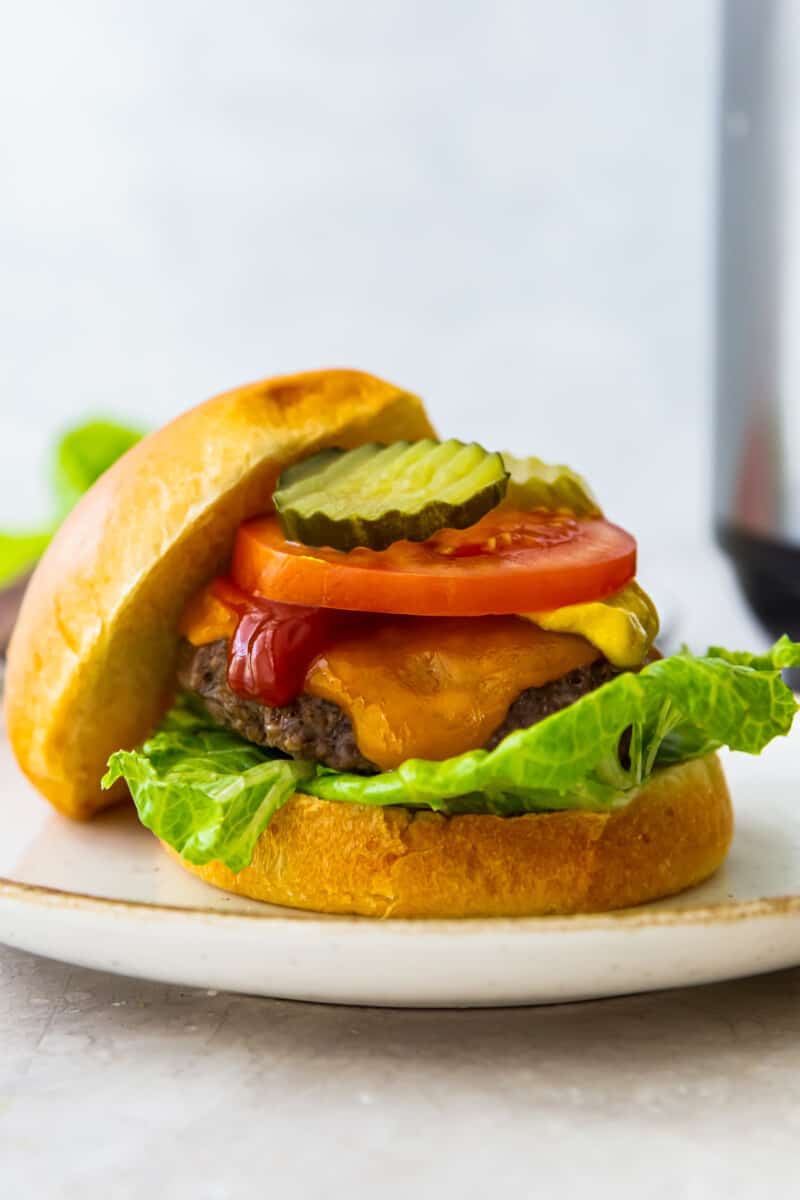 cheeseburger with a bun on a white plate topped with lettuce, cheese, ketchup, tomato slice, and pickles