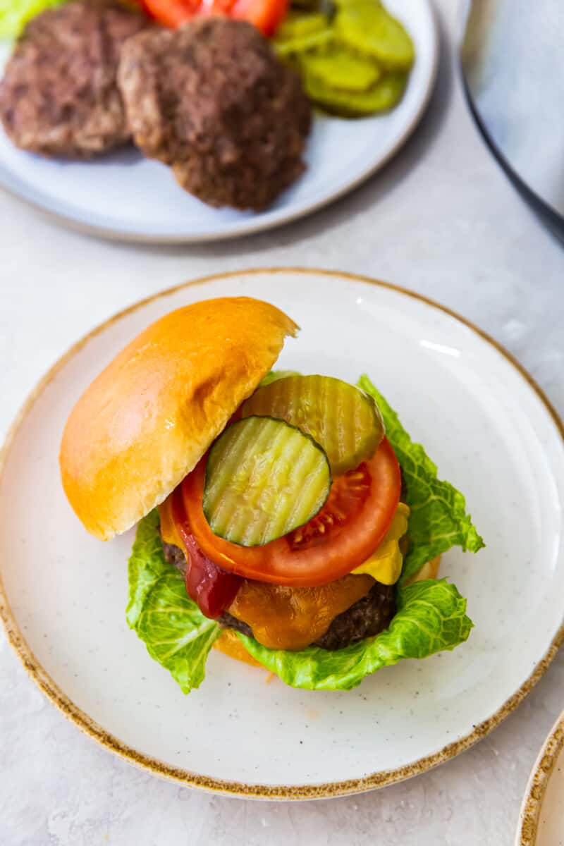 overhead image of cheeseburger with a bun on a white plate topped with lettuce, cheese, ketchup, tomato slice, and pickles