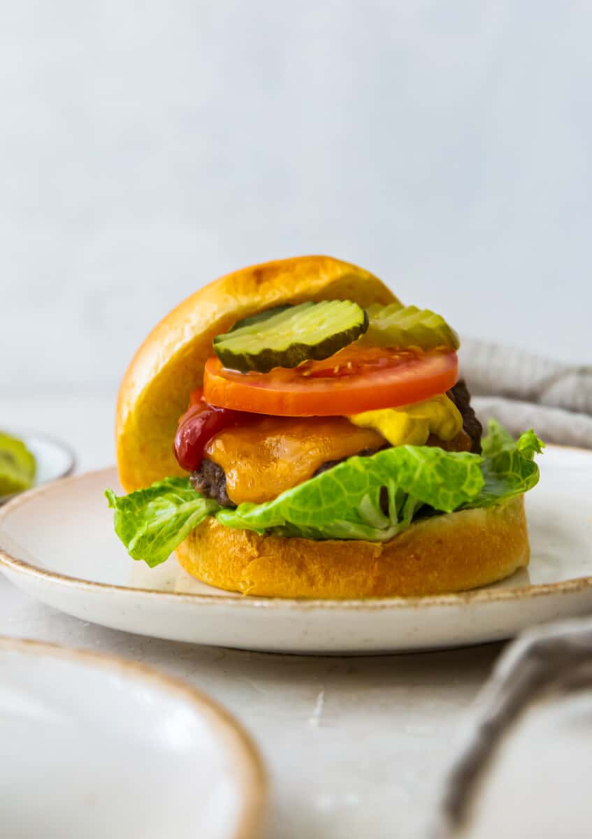 cheeseburger with a bun on a white plate topped with lettuce, cheese, ketchup, tomato slice, and pickles