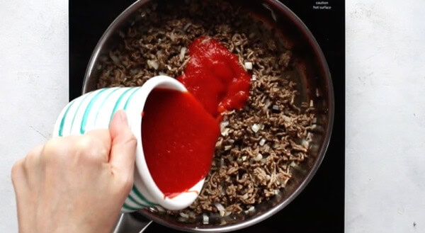 pouring marinara sauce into saucepan with ground beef, onions, and garlic