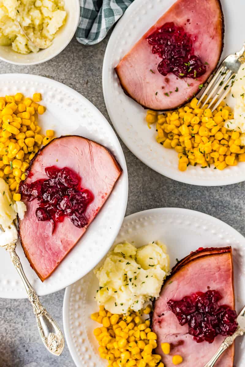 3 plates with slices of cranberry glazed ham, mashed potatoes, and corn