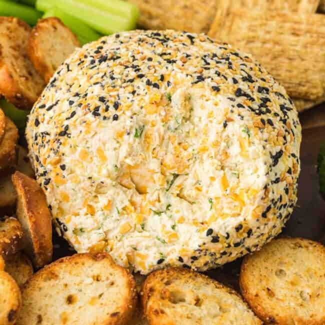 everything bagel cheeseball on a serving board with crackers, sliced cucumbers, and celery sticks with some of the cheeseball gone