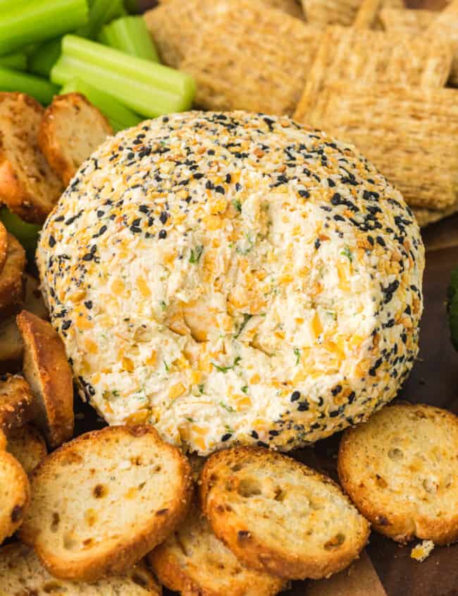 everything bagel cheeseball on a serving board with crackers, sliced cucumbers, and celery sticks with some of the cheeseball gone