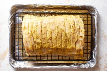 apple cider poured into the bottom of a rimmed baking sheet with a wire rack inset holding a mustard glazed raw pork tenderloin.