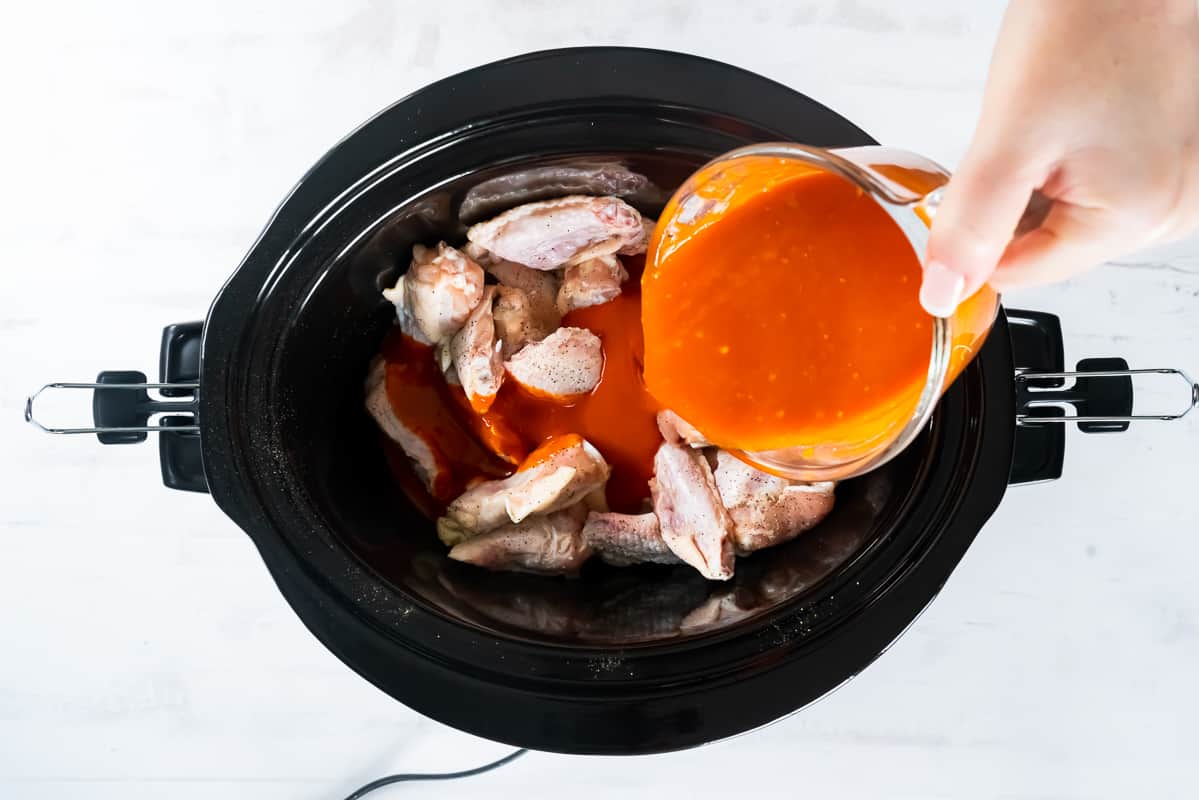 Pouring buffalo sauce into a Crock Pot filled with chicken wings.