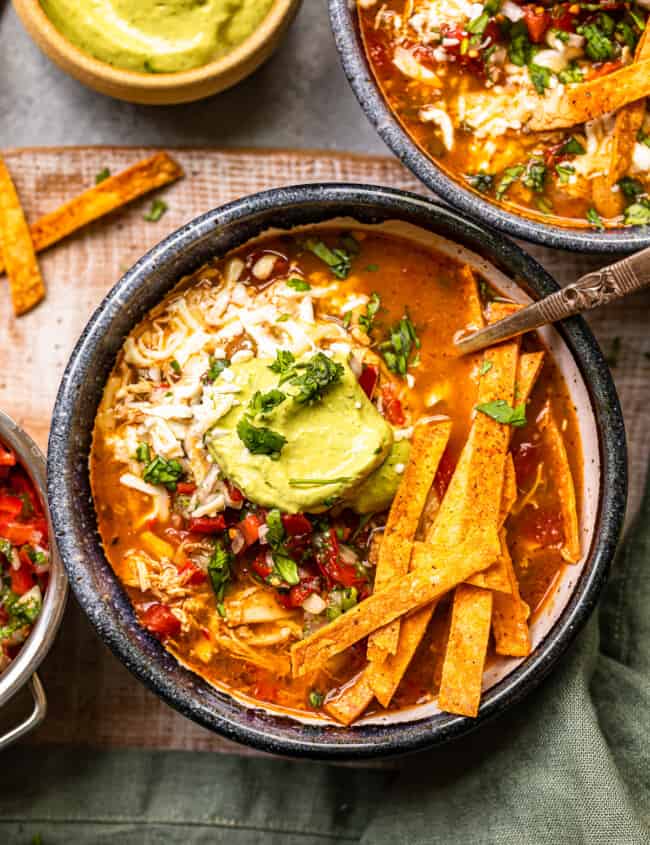 chicken fajita soup in a black bowl topped with cheese, guacamole, diced tomatoes, and tortilla strips with a spoon