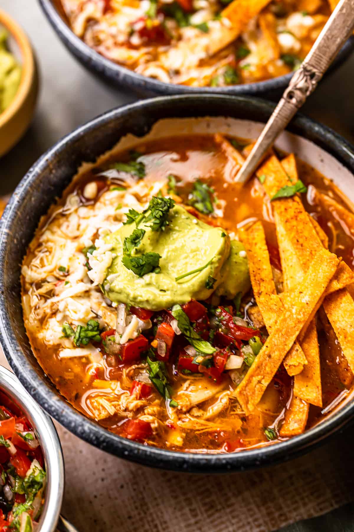 chicken fajita soup in a black bowl topped with cheese, guacamole, diced tomatoes, and tortilla strips with a spoon