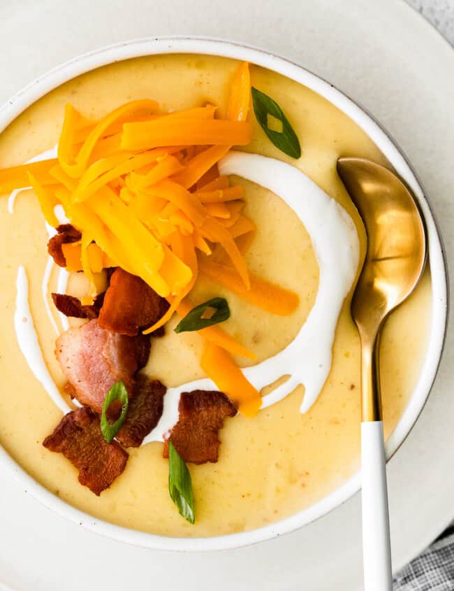 overhead image of creamy potato soup in a white bowl topped with shredded cheddar cheese and bacon with a spoon