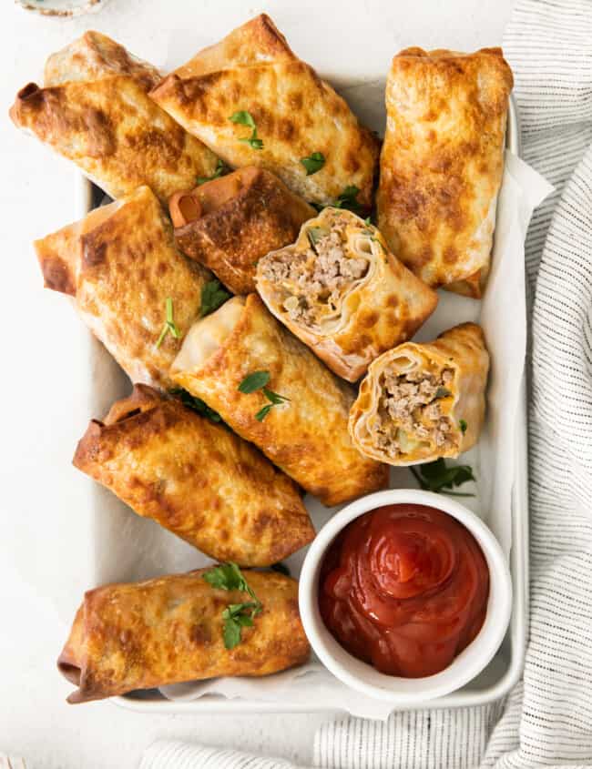 overhead image of cheeseburger egg rolls on a serving tray with a side of ketchup in a white bowl