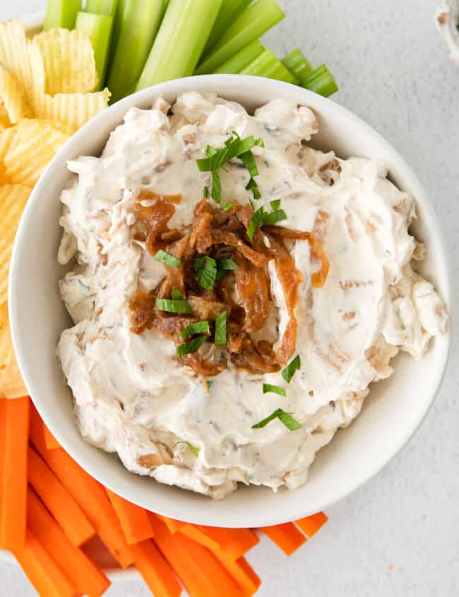 overhead image of french onion dip in a white bowl on a serving platter surrounded by chips, celery sticks, and carrot sticks