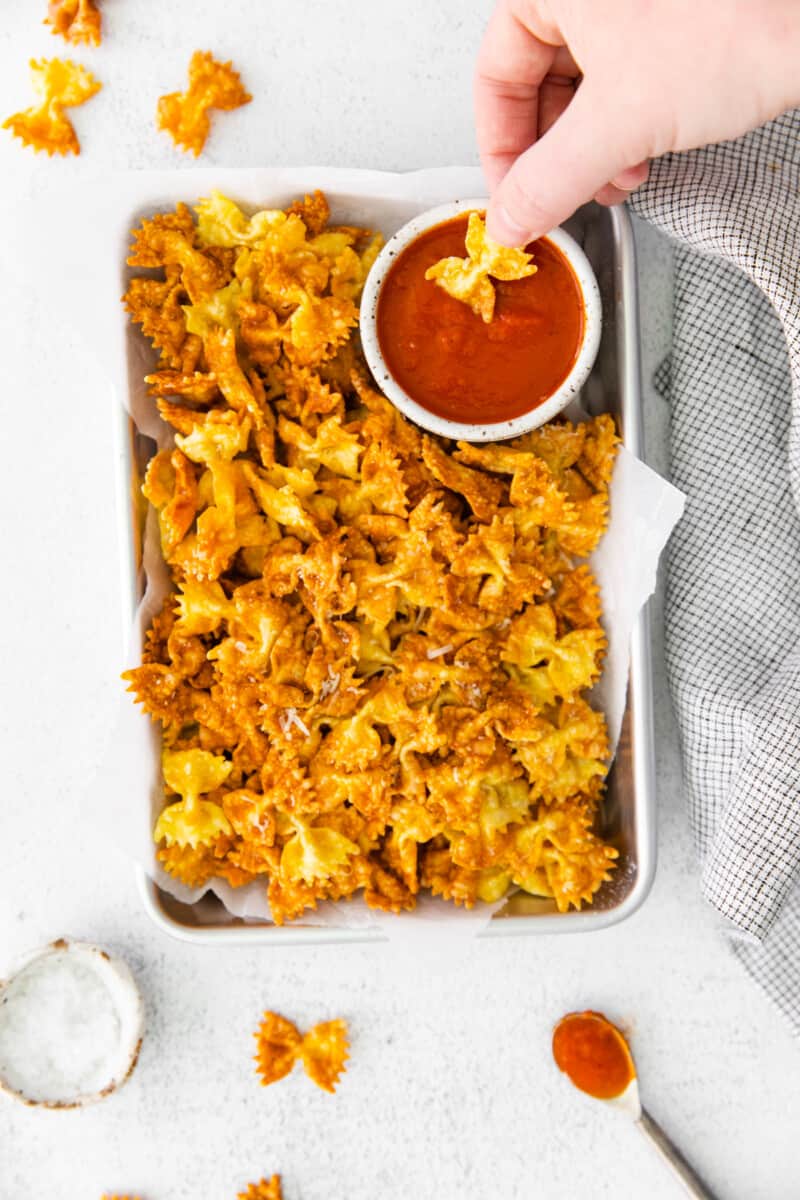 a hand dipping a garlic parmesan pasta chip into marinara sauce on a tray of pasta chips.