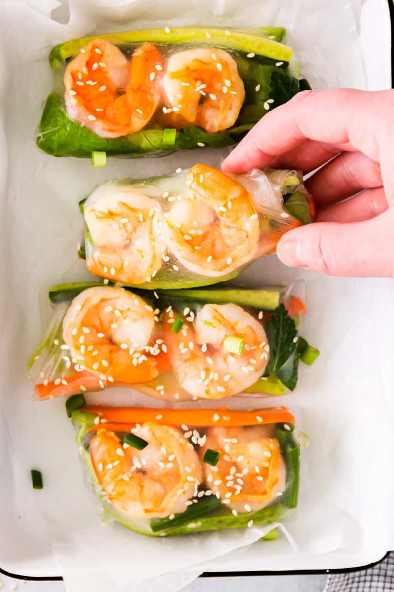 hand grabbing 1 of 4 shrimp summer rolls from a white rectangular tray.