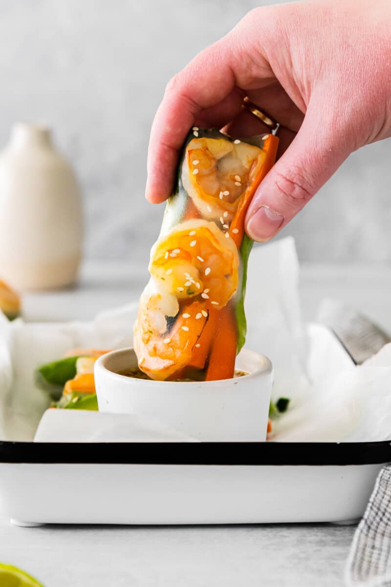 hand dipping 1 shrimp summer roll into dipping sauce in a white ramekin.