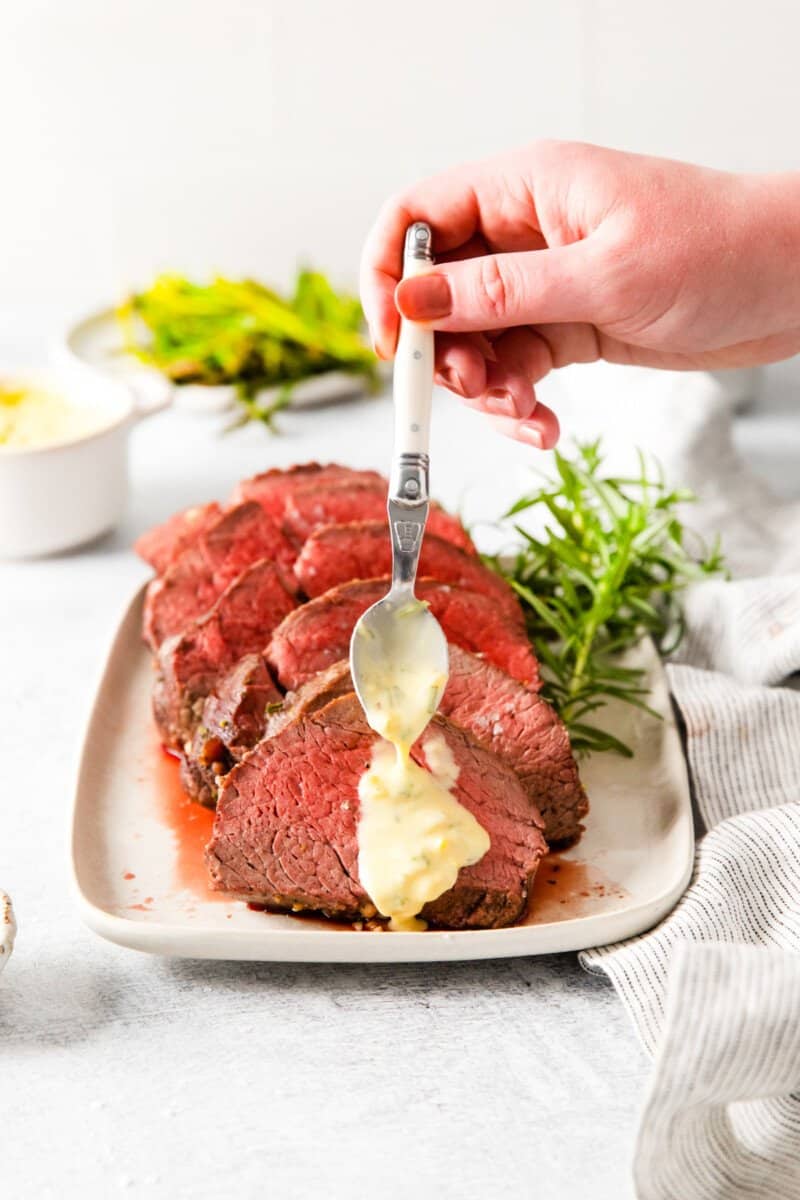 spoon pouring sauce over crockpot beef tenderloin on a white plate.