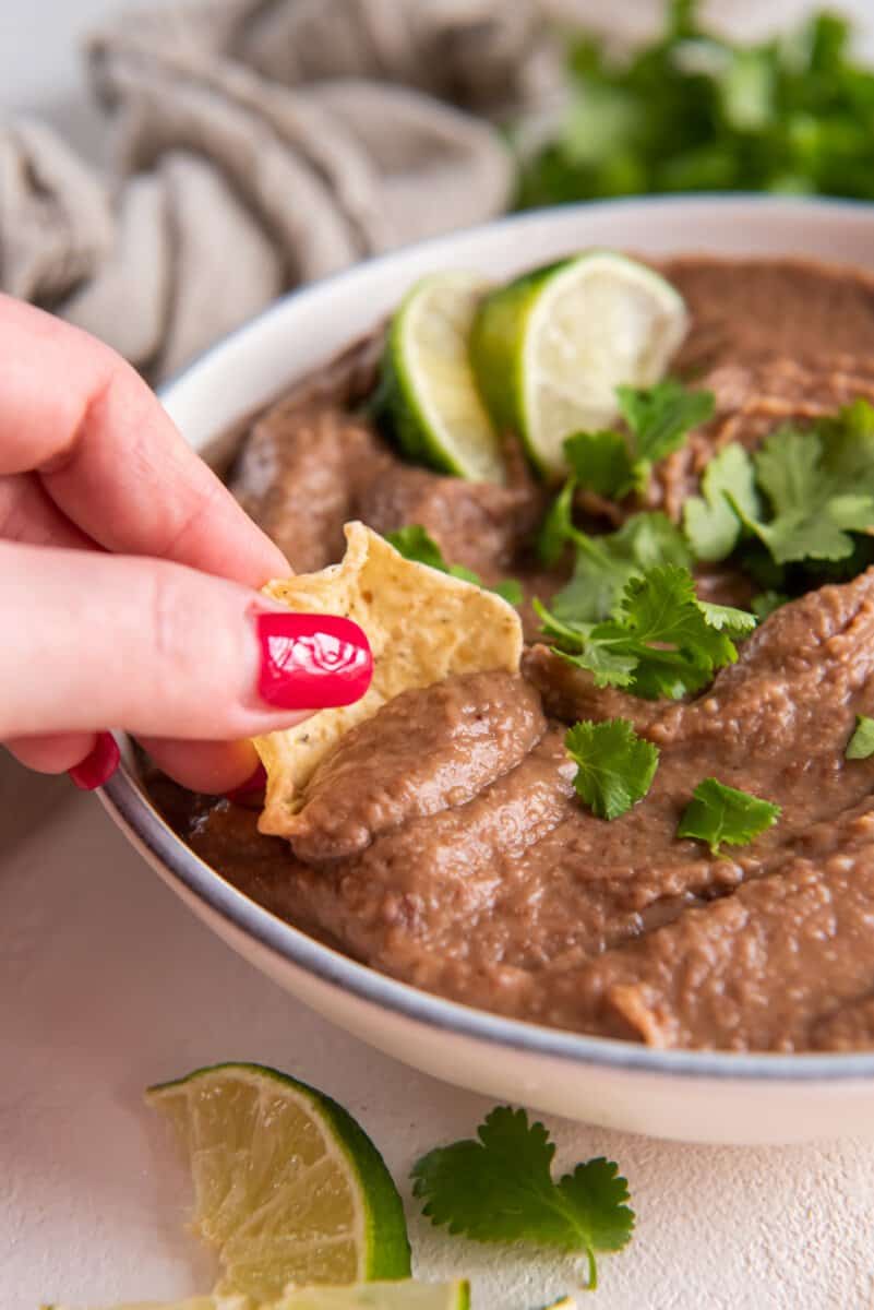 tortilla chip dipped into crockpot refried beans in a white bowl with cilantro and lime wedges.