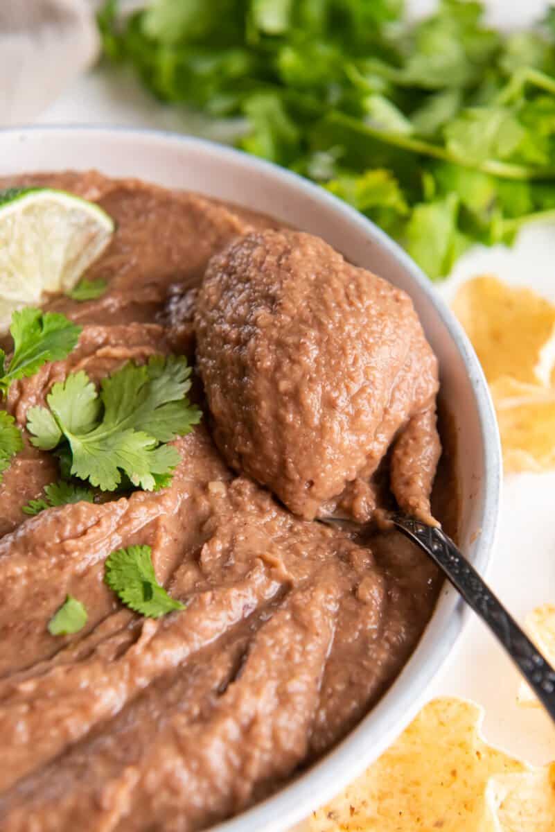 spoonful of crockpot refried beans resting on refried beans in a white bowl with cilantro and lime wedges.