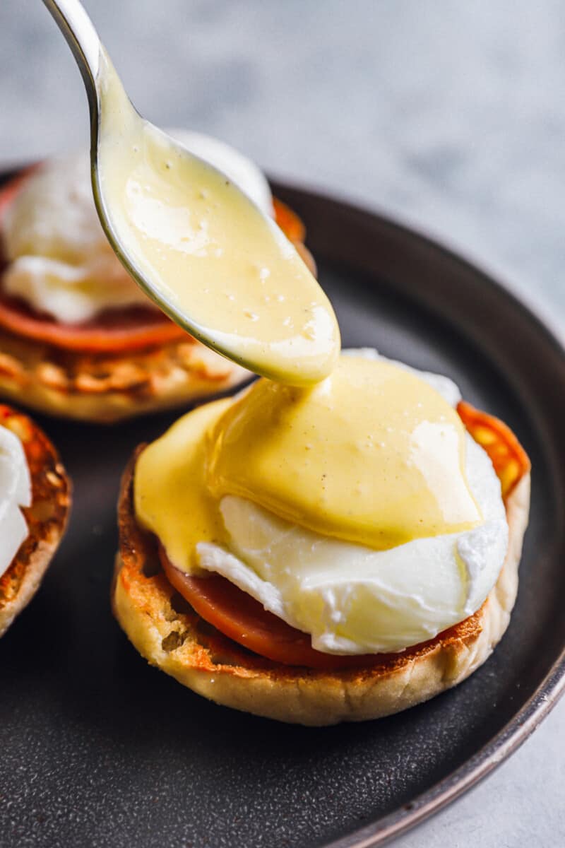 spoon pouring hollandaise sauce over eggs benedict on a black plate.