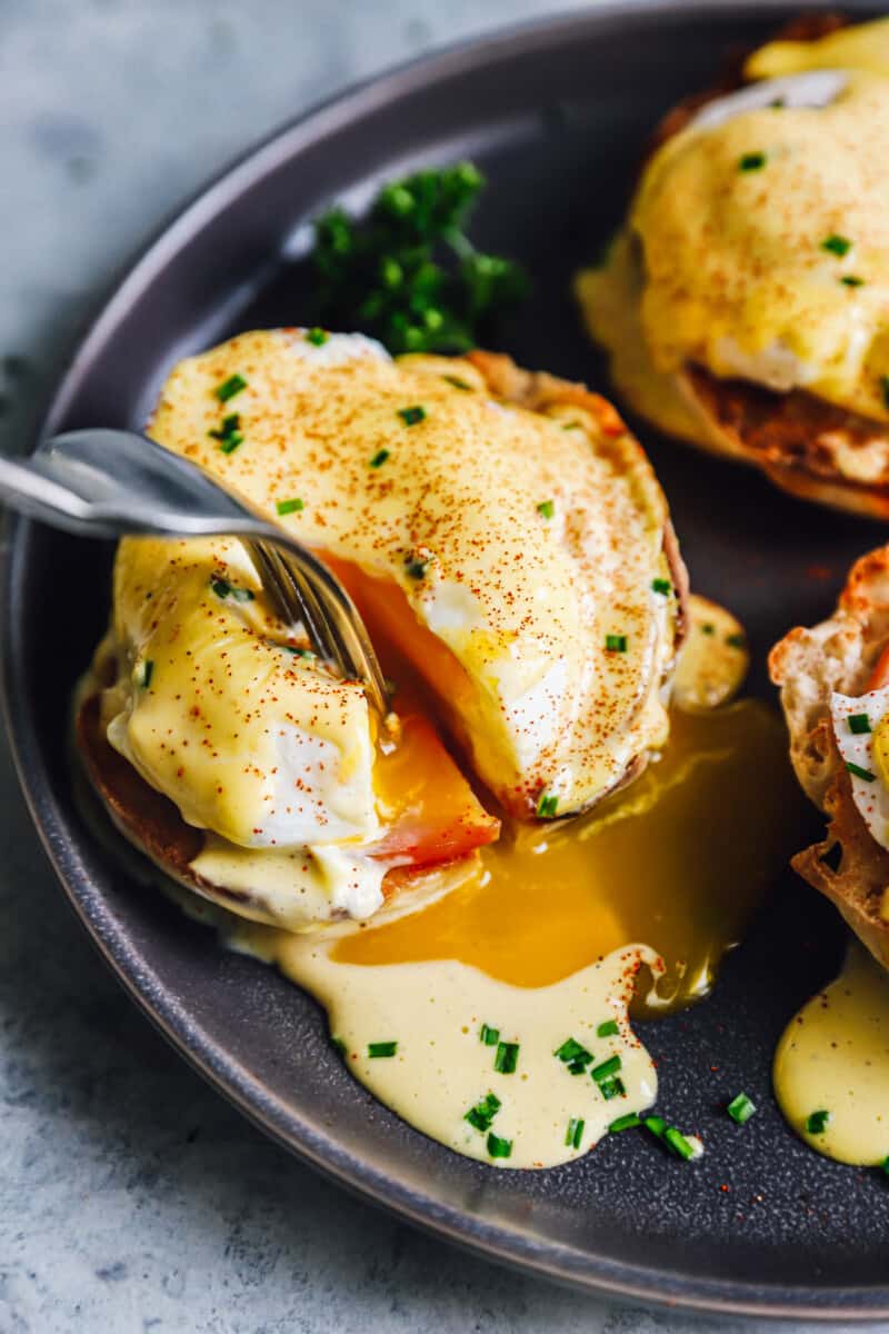 fork cutting open eggs benedict on a black plate.