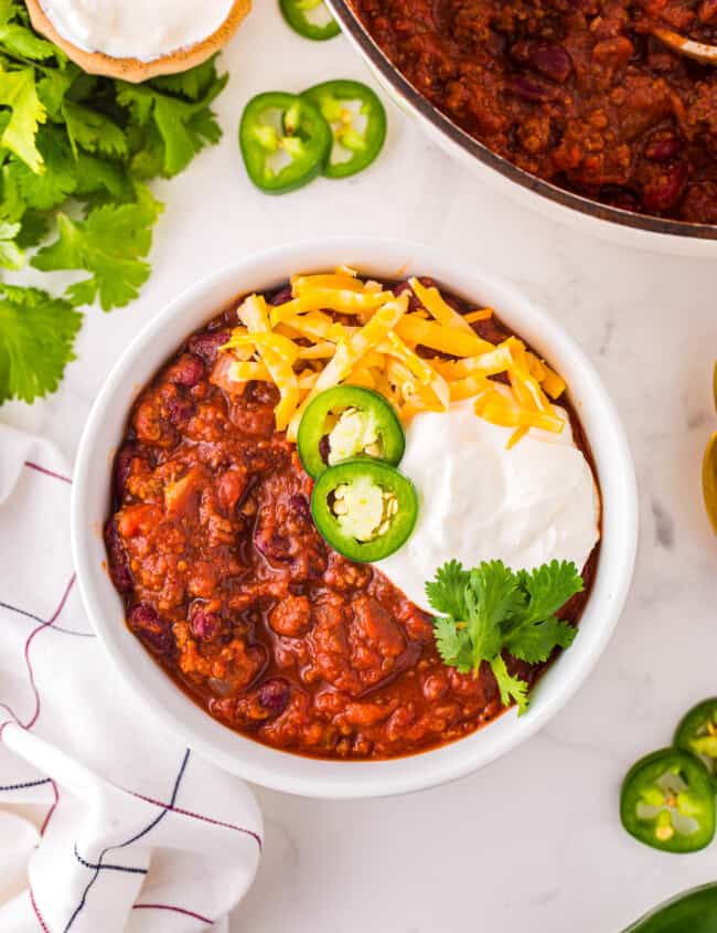 overhead view of chili con carne in a white bowl with cheese, sour cream, jalapenos, and cilantro.