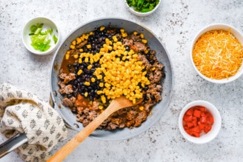 vegetables added to ground beef in a frying pan with a wooden spoon.