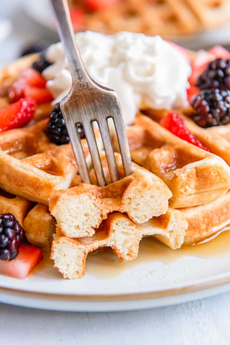 a forkful of belgian waffle in front of 2 stacked belgian waffles on a white plate with whipped cream and fruit.