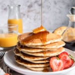 a tall stack of buttermilk pancakes on a white plate with butter, syrup, and a halved strawberry.