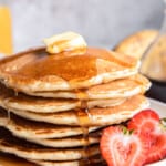 close up of a tall stack of buttermilk pancakes on a white plate with butter, syrup, and a halved strawberry.