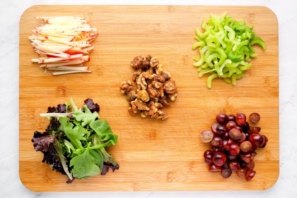 Waldorf salad ingredients grouped together on a cutting board (thinly sliced apples, halved grapes, candied walnuts, sliced celery, greens)